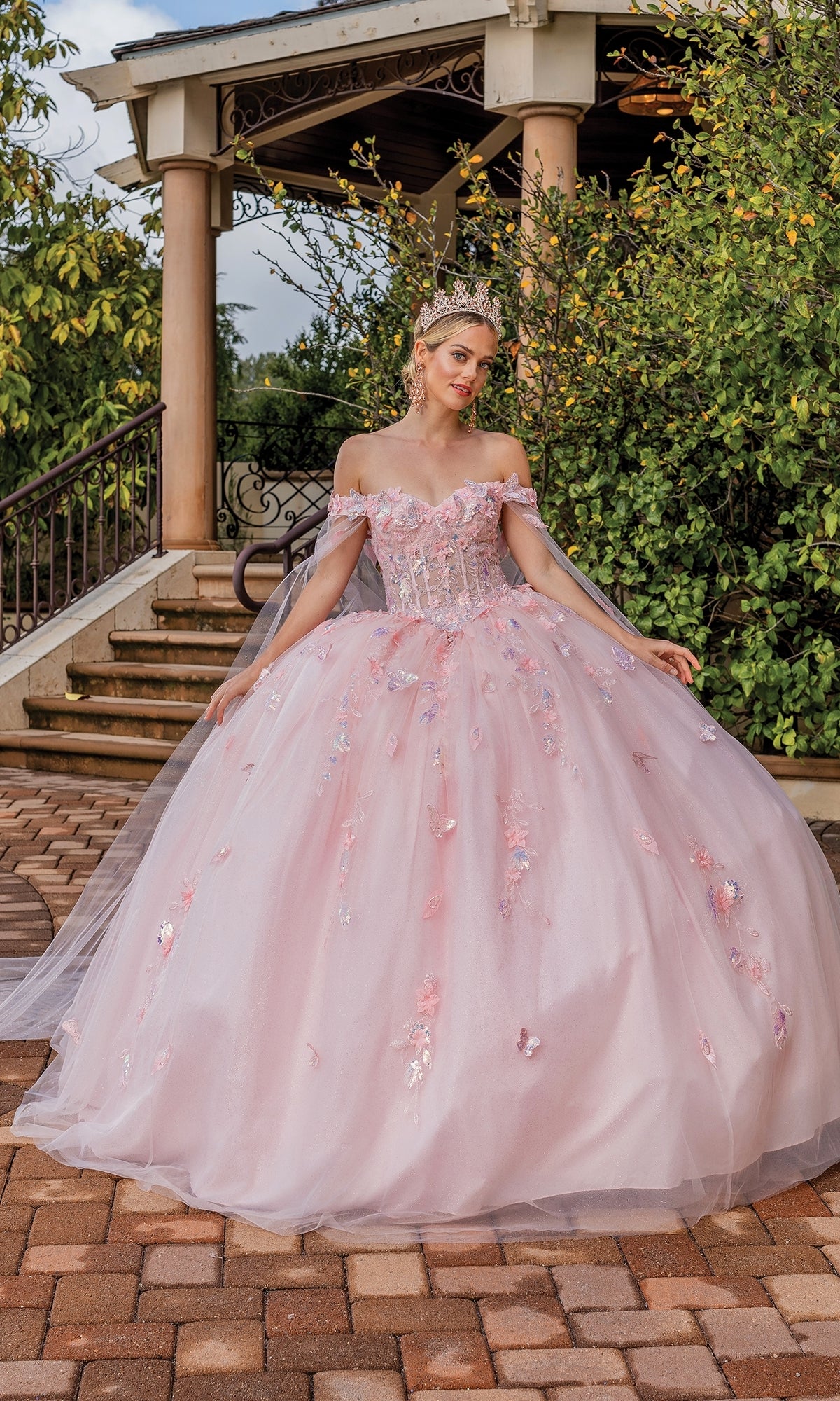 A woman in the Quinceanera Dress 1840 By Dancing Queen, a pink off-shoulder ball gown with floral details and a tiara, stands outdoors before a garden gazebo.